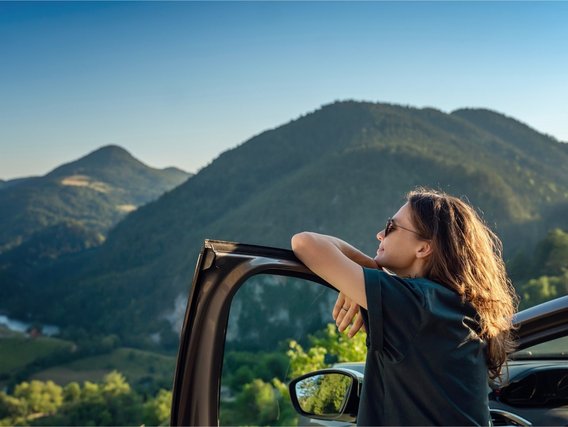 MPU-Vorbereitung mit TÜV NORD-KURS Frau lehnt entspannt an Autotür und blickt in bergige Landschaft