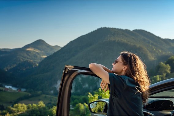 Frau lehnt entspannt an Autotür und blickt in bergige Landschaft