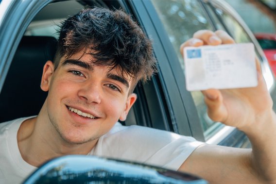 Junger Mann sitzt im Auto und hält lächelnd seinen Führerschein aus dem Fenster