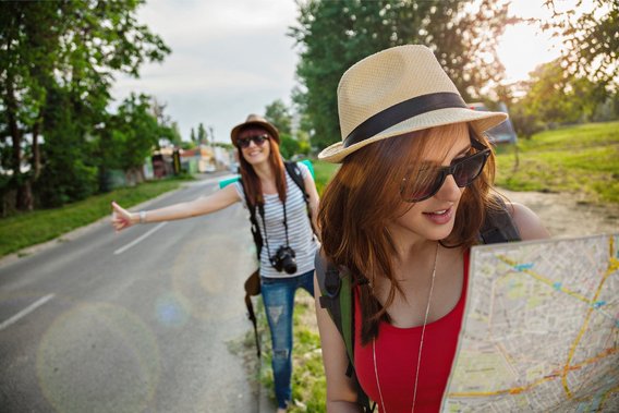 Zwei junge Frauen auf Landstraße mit Karte und Daumen raus – auf Reisen ohne Führerschein