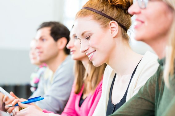 Lächelnde junge Frau sitzt mit Stift im Seminar neben weiteren Teilnehmenden