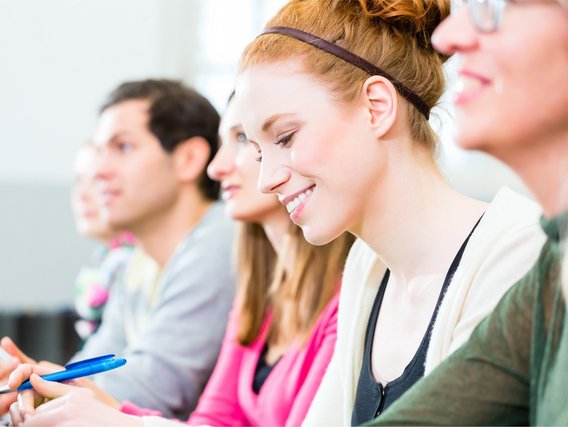 Lächelnde junge Frau sitzt mit Stift im Seminar neben weiteren Teilnehmenden